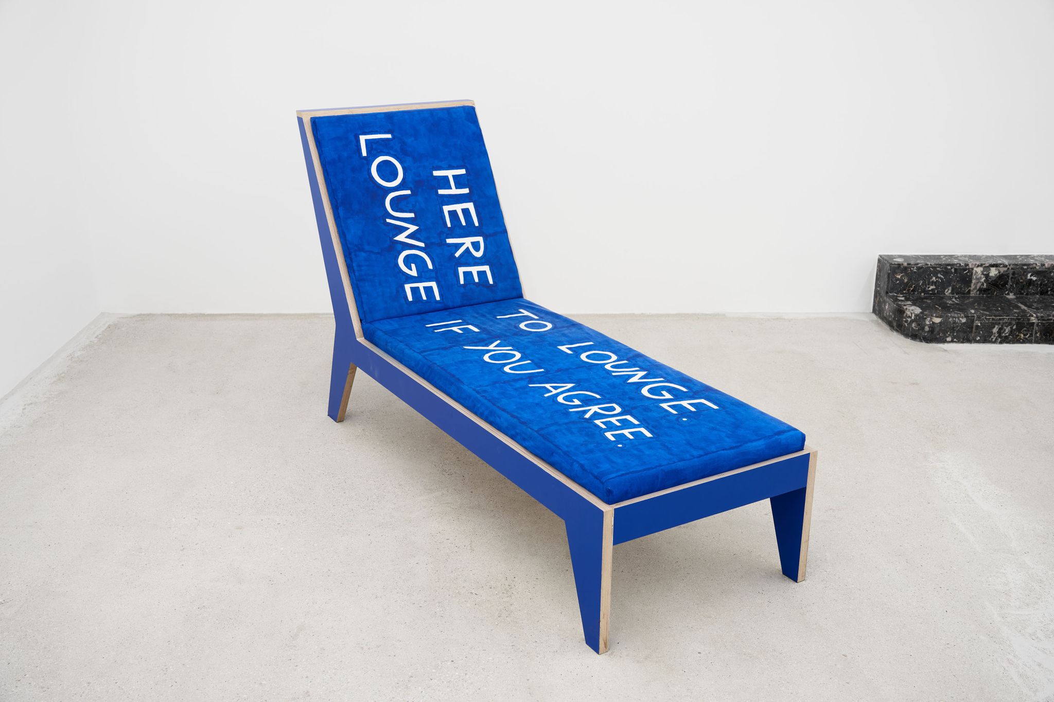 Finnegan Shannon, Do you want us here or not (MMK) – Chaise lounge 3, 2021, Plywood, paint, foam, fabric, fabric paint, 110.5 ⁠× ⁠190 ⁠× ⁠68.5 ⁠⁠cm, Image description: A blue chaise lounge is standing in an exhibition room. On it, white letters read “Here to lounge. Lounge if you agree”.