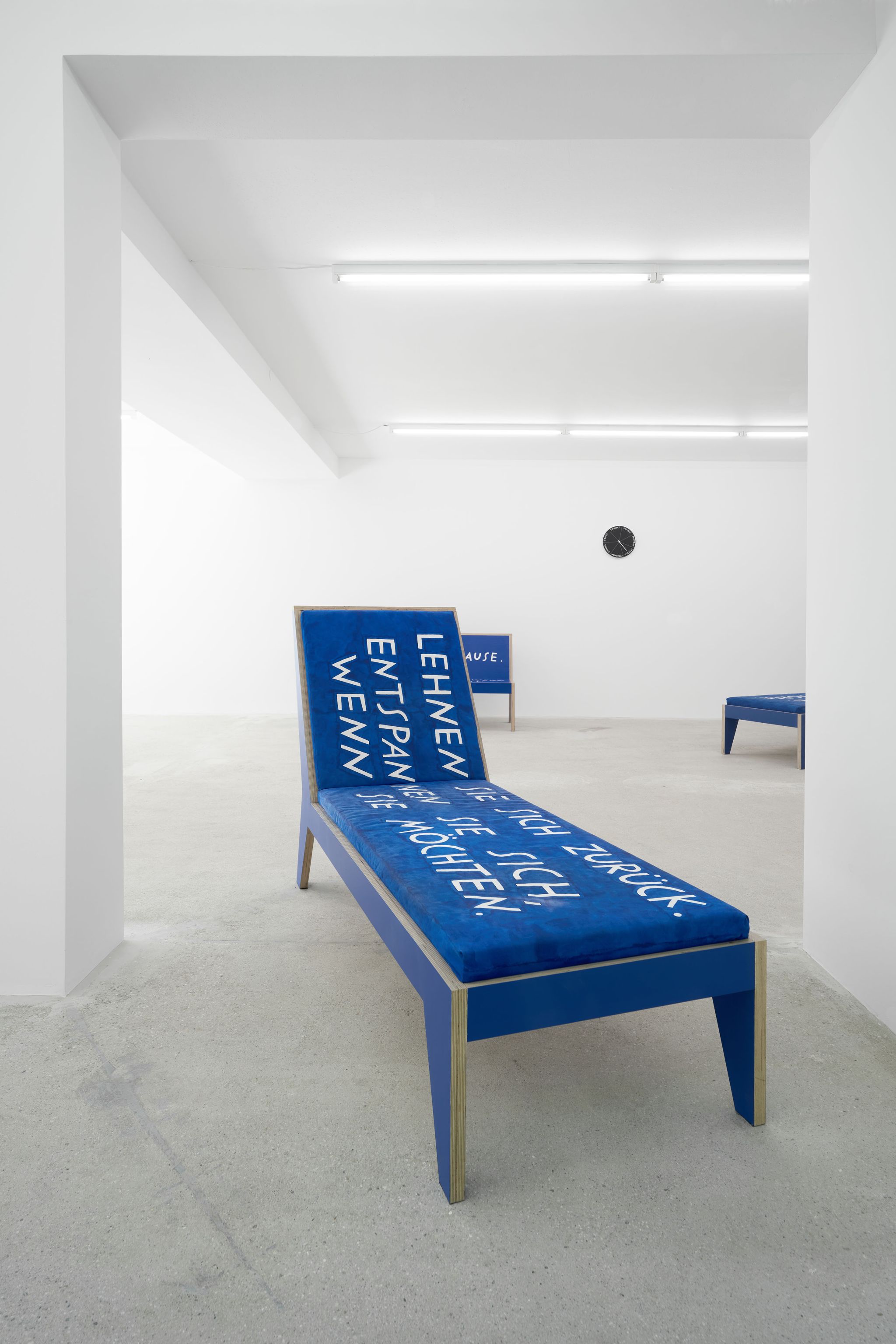Finnegan Shannon, Do you want us here or not (MMK) – Chaise lounge 1, 2021, Plywood, paint, foam, fabric, fabric paint, 110.5 ⁠× ⁠190 ⁠× ⁠68.5 ⁠⁠cm, Image description: Two blue chaise lounges and a blue bench are in an exhibition room. All have different hand-painted text on them. A black clock is hanging on the wall in the background.