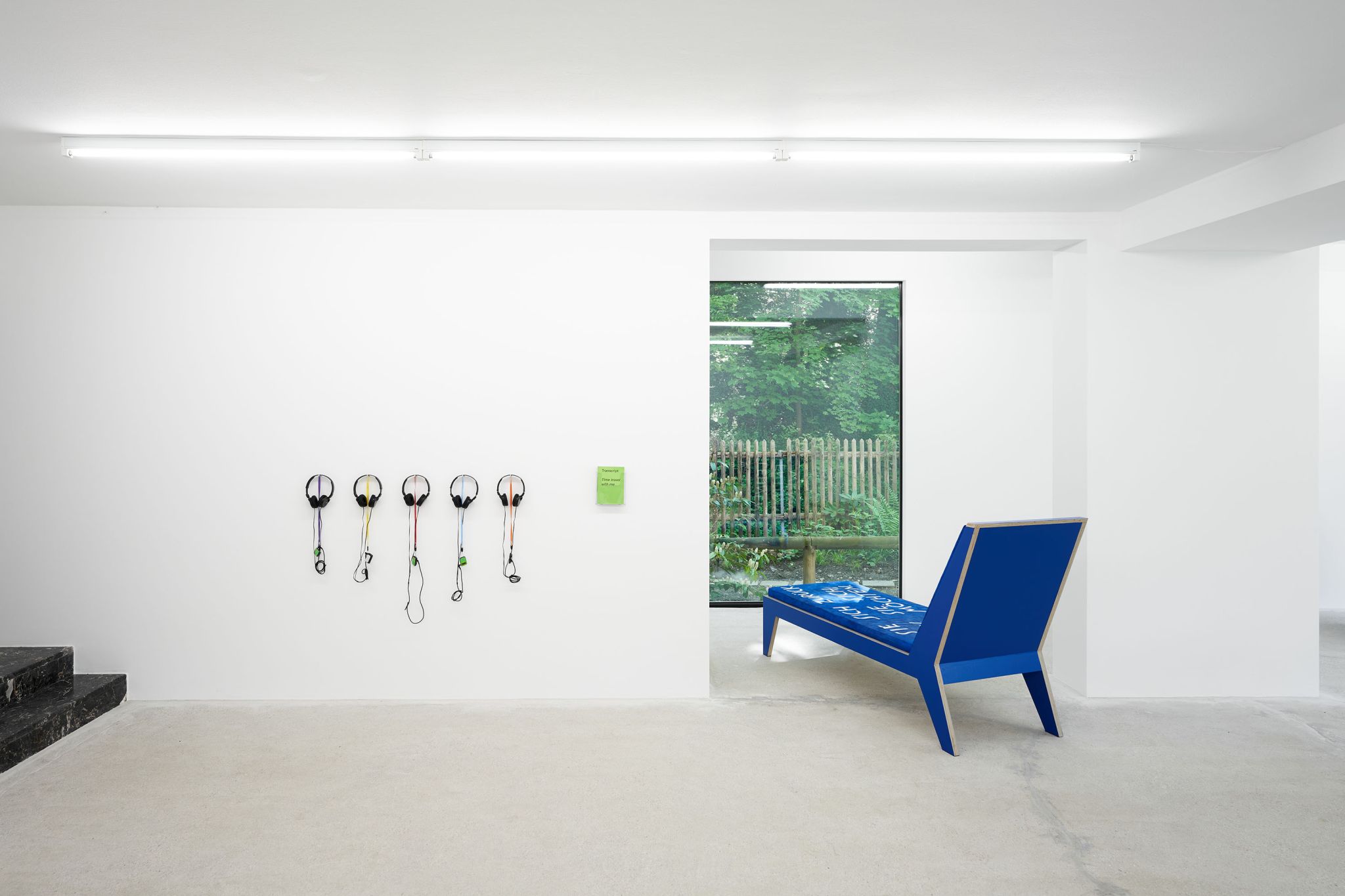 Finnegan Shannon, <em>Slower,</em> Installation view, Deborah Schamoni, 2022, Image description: Five headphones plugged into mp3 players are hanging on a wall next to a blue chaise lounge facing out of the window.