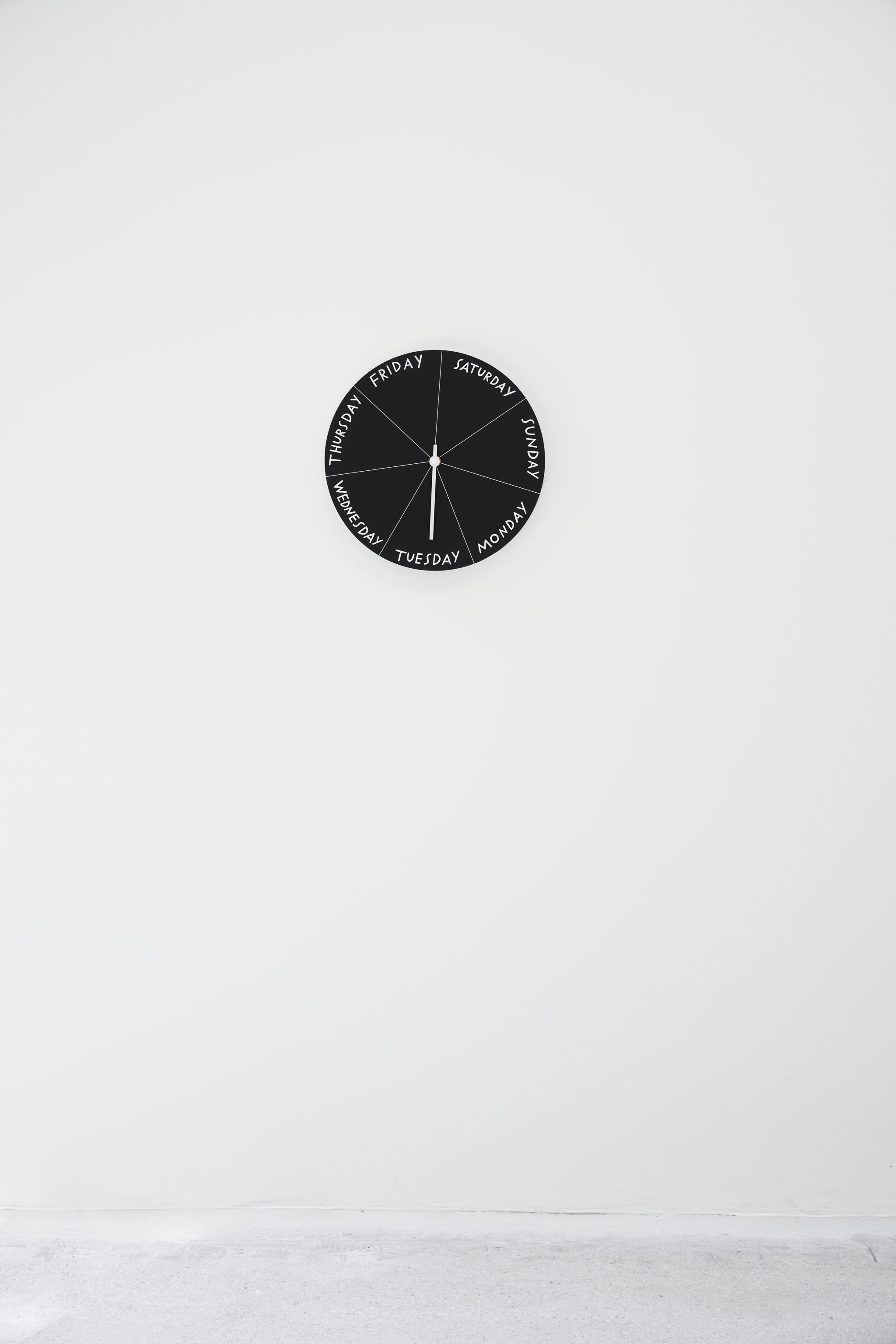 Finnegan Shannon, Have you ever fallen in love with a clock? – Saturday, 2021, Day Clock mechanism, DiBond, clock hand, paint, ø 34 ⁠cm, Image description: A black Day Clock is hanging on a wall, the white clock hand is pointing to Tuesday. On the 1 o’clock position, it says “Saturday”. The other days of the week follow around the clock.