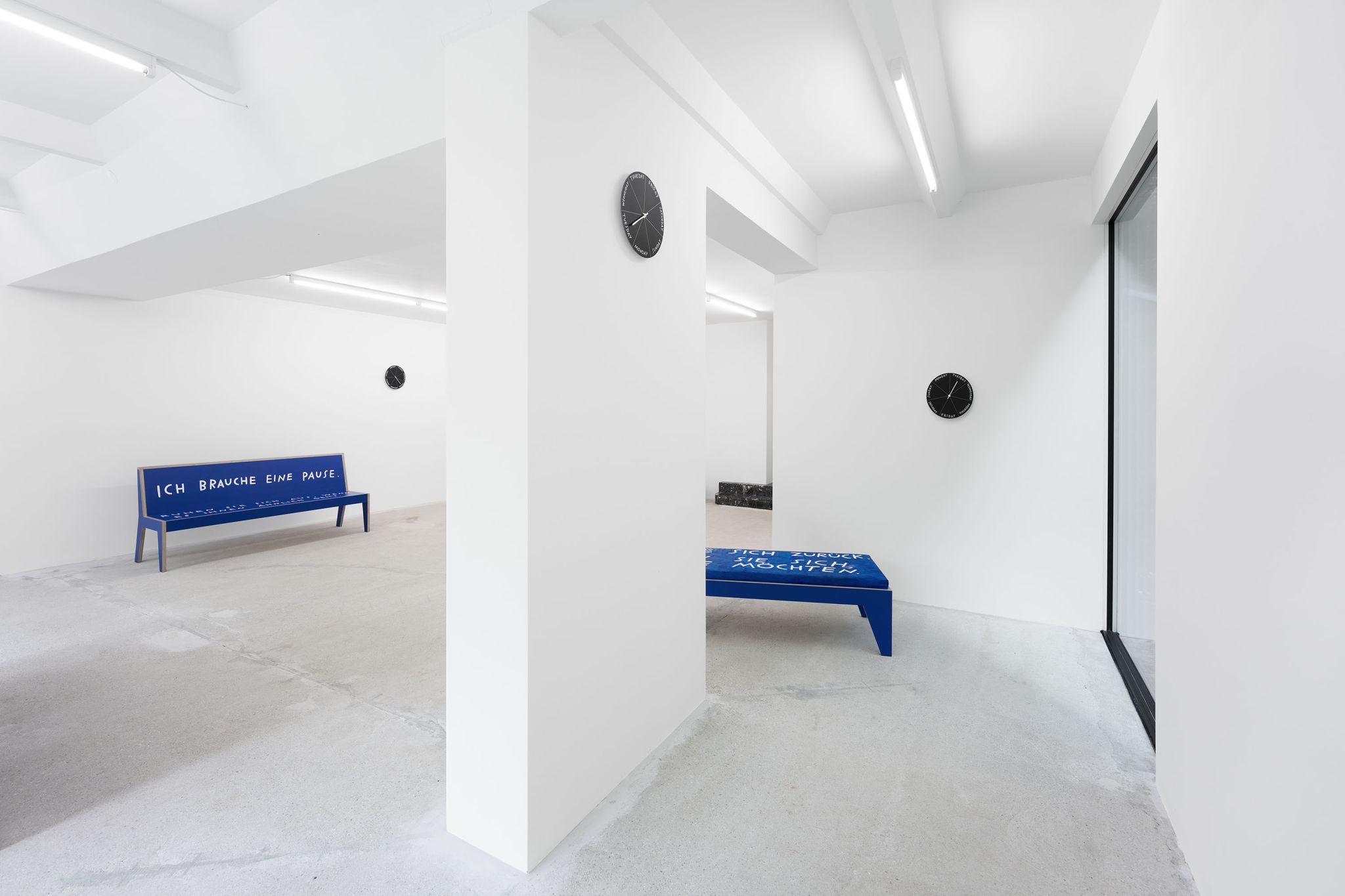 Finnegan Shannon, <em>Slower,</em> Installation view, Deborah Schamoni, 2022, Image description: Inside an exhibition room there is one blue bench with white letters on it. Part of another piece of blue furniture is in view, cut off by column in the space. Three black clocks are hanging on different walls at different heights.