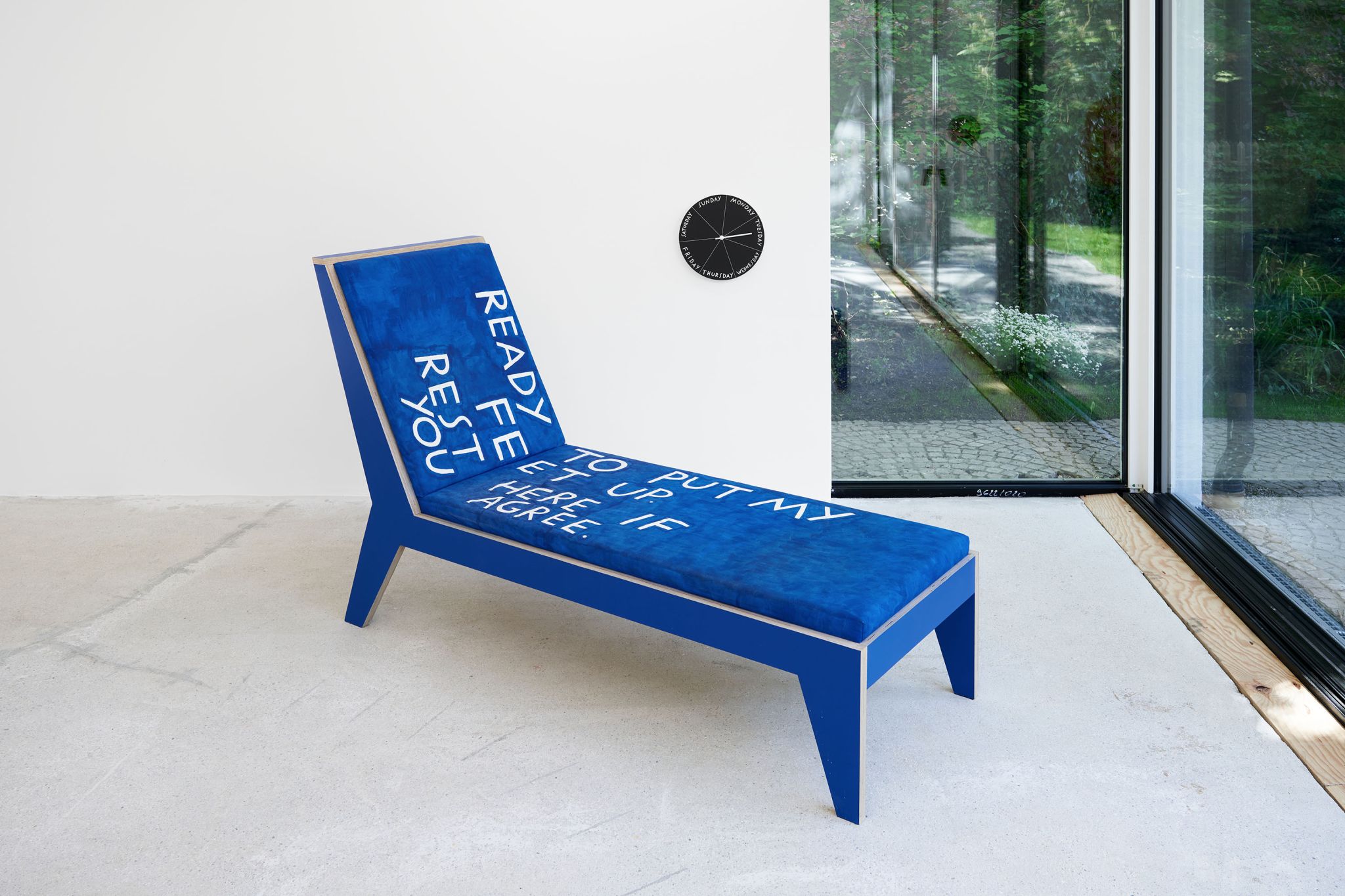 Finnegan Shannon, Do you want us here or not (MMK) – Chaise lounge 2, 2021, Plywood, paint, foam, fabric, fabric paint, 110.5 ⁠× ⁠190 ⁠× ⁠68.5 ⁠⁠cm, Image description: A blue chaise lounge with white letters on it in an exhibition room. The letters read “Ready to put my feet up. Rest here if you agree.” The chaise lounge is facing out of the big window with views of a lush garden. A black Day Clock is hanging next to it.
