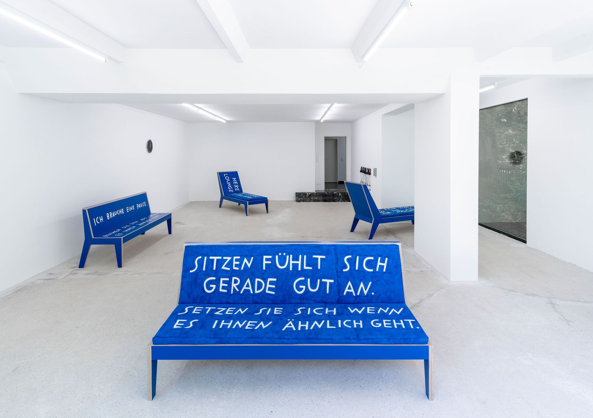 Finnegan Shannon, <em>Slower,</em> Installation view, Deborah Schamoni, 2022, Image description: Two blue benches and two chaise lounges in an exhibition room, all have text written in white letters on them. They are all facing different directions, scattered around the room. In the background, there’s a black clock and on the opposite side of the room, there are five headphones hanging on a wall.