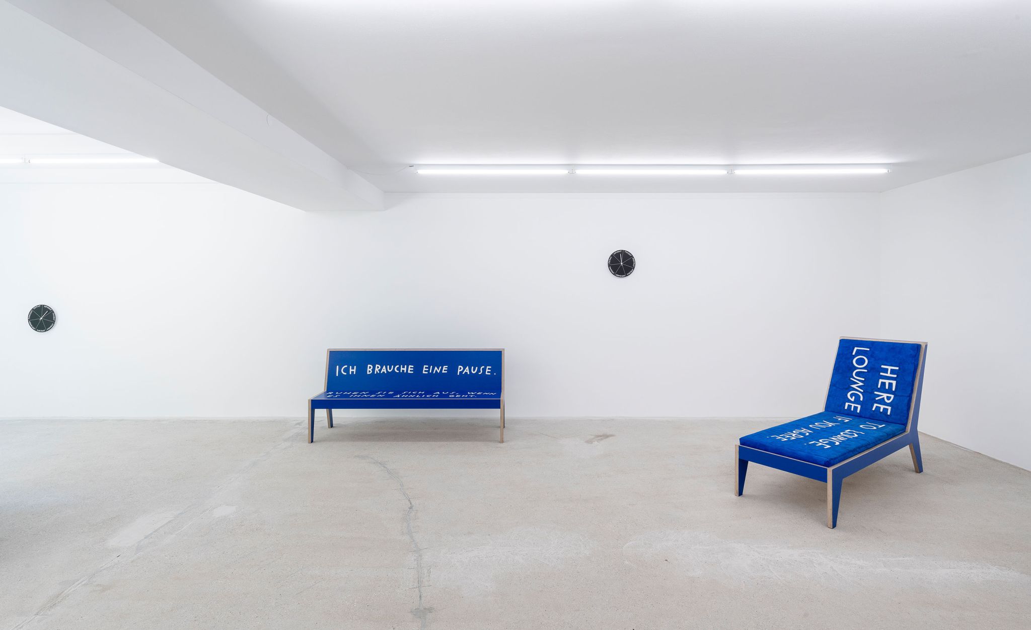 Finnegan Shannon, <em>Slower,</em> Installation view, Deborah Schamoni, 2022, Image desription: A blue bench and a blue chaise lounge in an exhibition room. Both have text in white letters on them, one in German, the other in English language. Both texts invite the viewer to rest. Two black clocks are hanging on the wall in different heights.