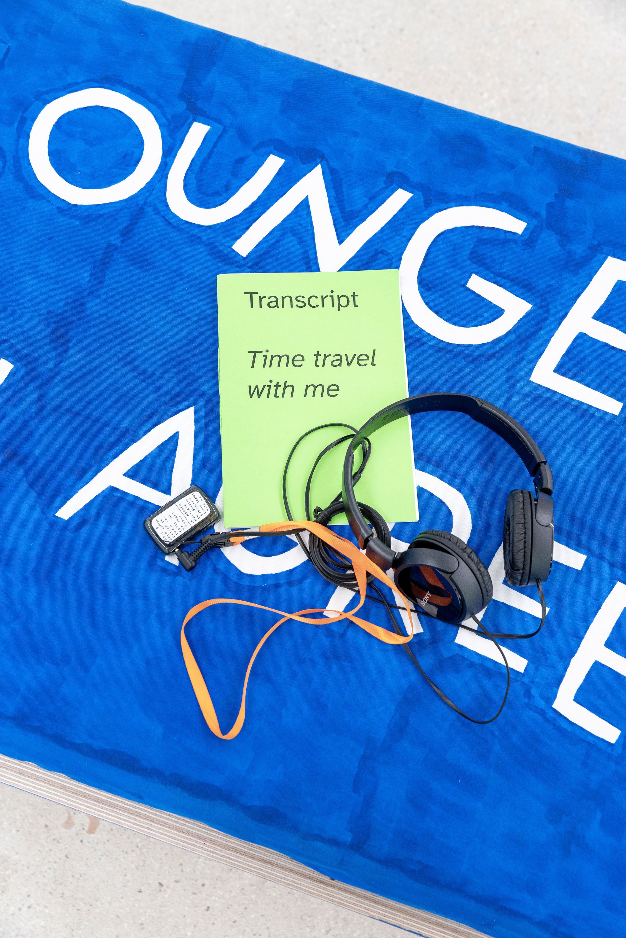 Finnegan Shannon, Time travel with me, 2022, Audio, mp3-Player, lanyard, headphones, transcript, zine, Image description: Headphones plugged into an mp3-Player and a zine laying on a blue piece of furniture. On the zine, it says “Transcript” and “Time travel with me.” The player is flipped, the back shows a sticker with handwritten instructions for how to play the audio.