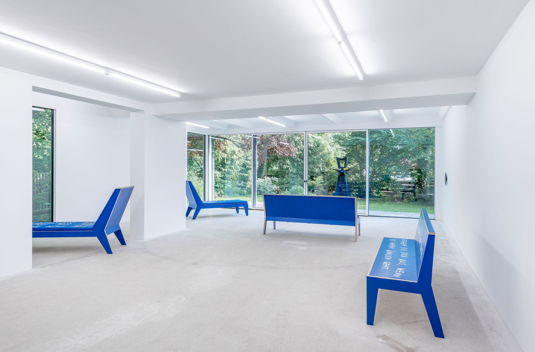 Finnegan Shannon, <em>Slower,</em> Installation view, Deborah Schamoni, 2022, Image description: Two blue chaise lounges and two blue benches in an exhibition room. They all face towards different windows into a garden.