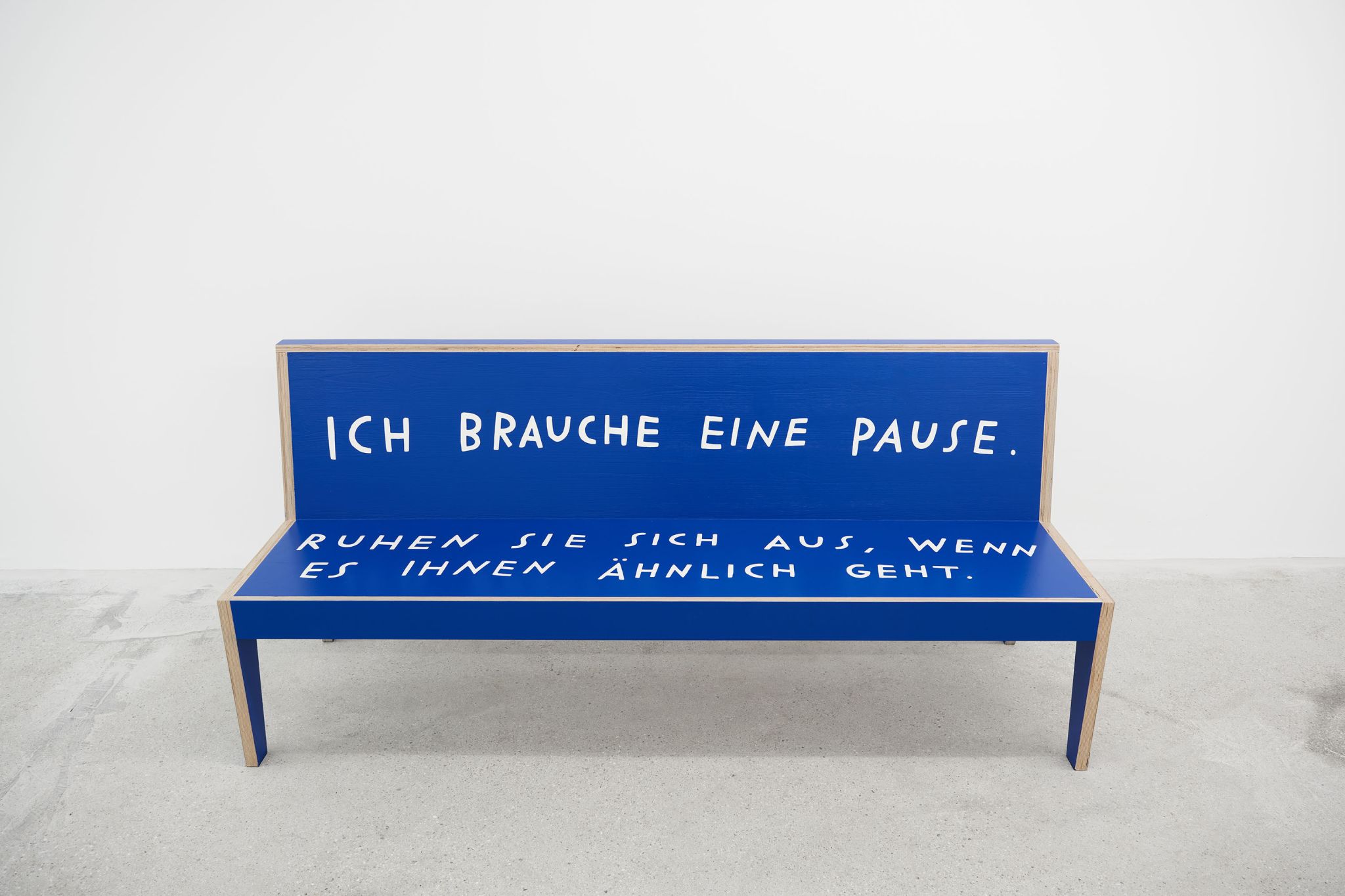 Finnegan Shannon, o you want us here or not (MMK) – Bench 6, 2021, Plywood, paint, 90 ⁠× ⁠200 ⁠× ⁠55 ⁠⁠cm, Image description: A blue bench is standing in an exhibition room. It has white letters on it, that read “I need a break. Rest here if you agree” in German language.