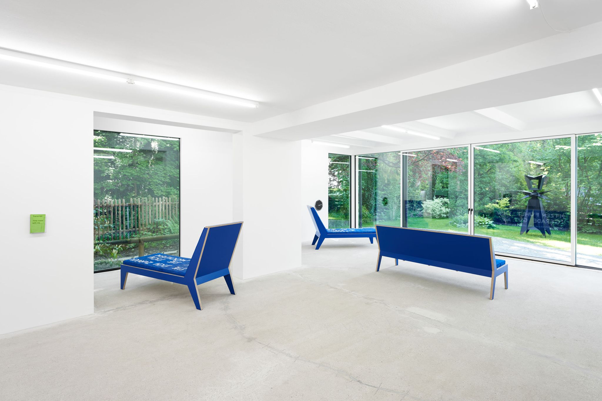 Finnegan Shannon, <em>Slower,</em> Installation view, Deborah Schamoni, 2022, Image description: Two blue chaise lounges and a blue bench in an exhibition room. They all face towards different windows into a garden. A green zine and a black clock are hanging on different walls.