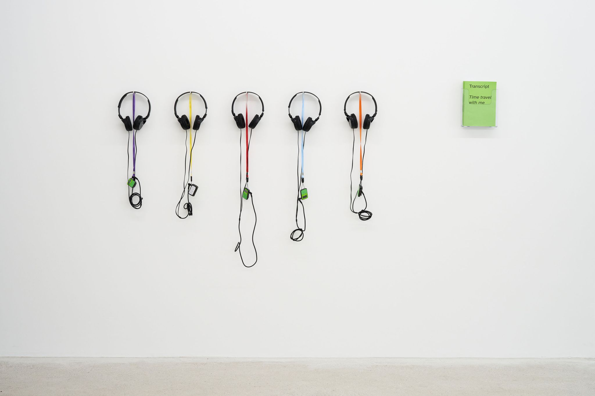Finnegan Shannon, Time travel with me, 2022, Audio, mp3-Player, lanyard, headphones, transcript, zine, Image description: Five headphones plugged into mp3 players are hanging on a wall. Next to them a zine with a bright green cover. On it, it says “Transcript” and “Time travel with me.”